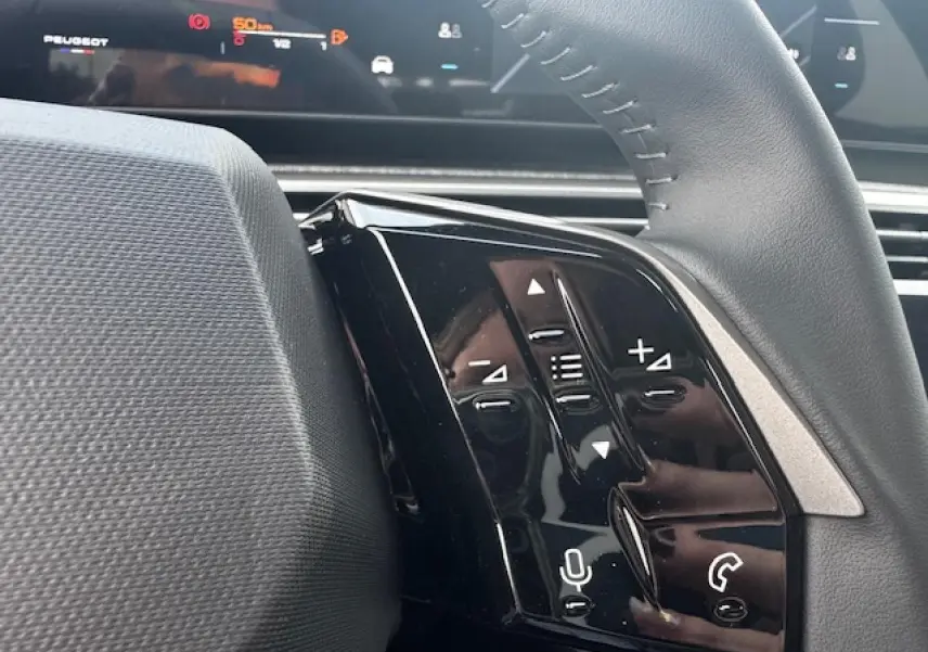 Gros plan sur les commandes noires du volant côté gauche du Peugeot 5008 2025, avec affichage numérique en arrière-plan.