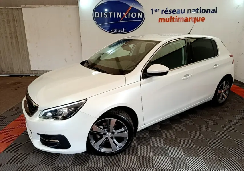 Peugeot 308 blanc vue 3/4 avant droit en intérieur, avec jantes alliage et phares ellipsoïdaux visibles.