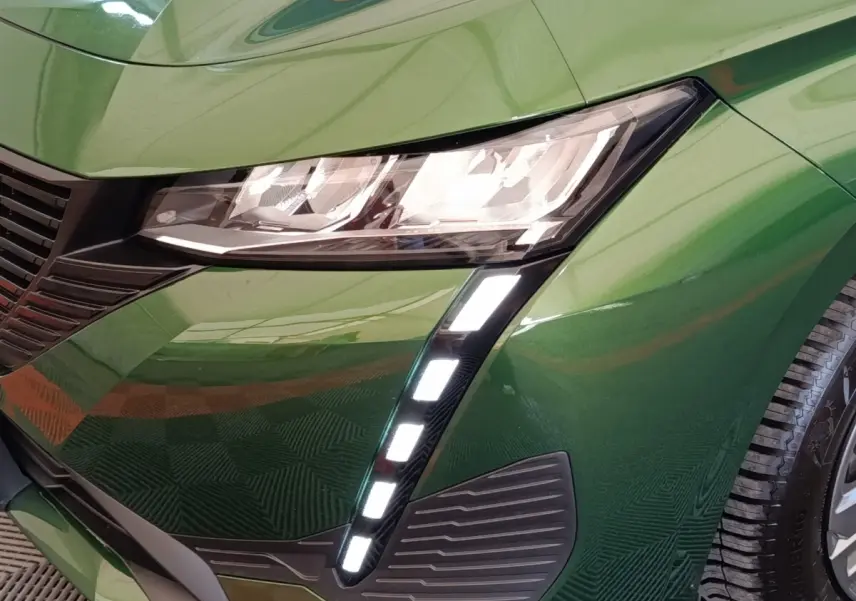 Gros plan sur l'avant droit vert métallisé d'une Peugeot 308 2023, mettant en valeur les phares LED et la calandre noire.