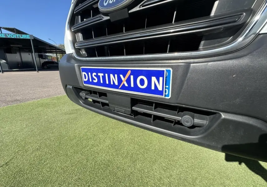 Gros plan sur la calandre avant noire et la plaque "DISTINXION" du Ford Transit Fourgon blanc de 2020.