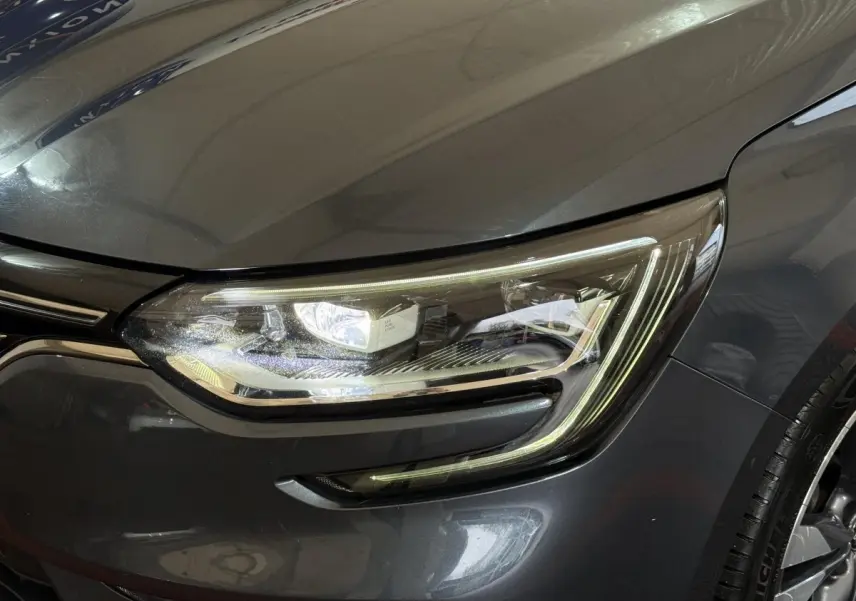 Gros plan sur le phare avant droit d’une Renault Mégane Intens gris foncé, avec détails LED et partie de la calandre visible.