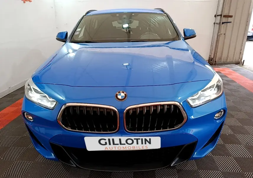 Vue avant d'un BMW X2 sDrive20i M Sport bleu, avec calandre noire et phares LED allumés, en intérieur showroom.