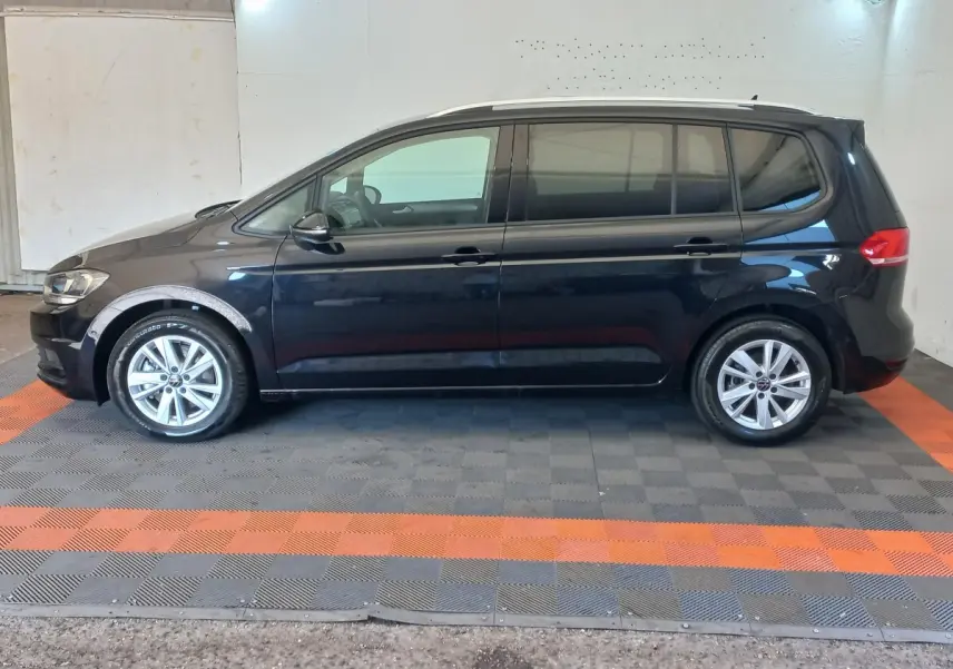 Vue de profil côté gauche d’un Volkswagen Touran noir 7 places, sur sol à damier gris et orange en intérieur.