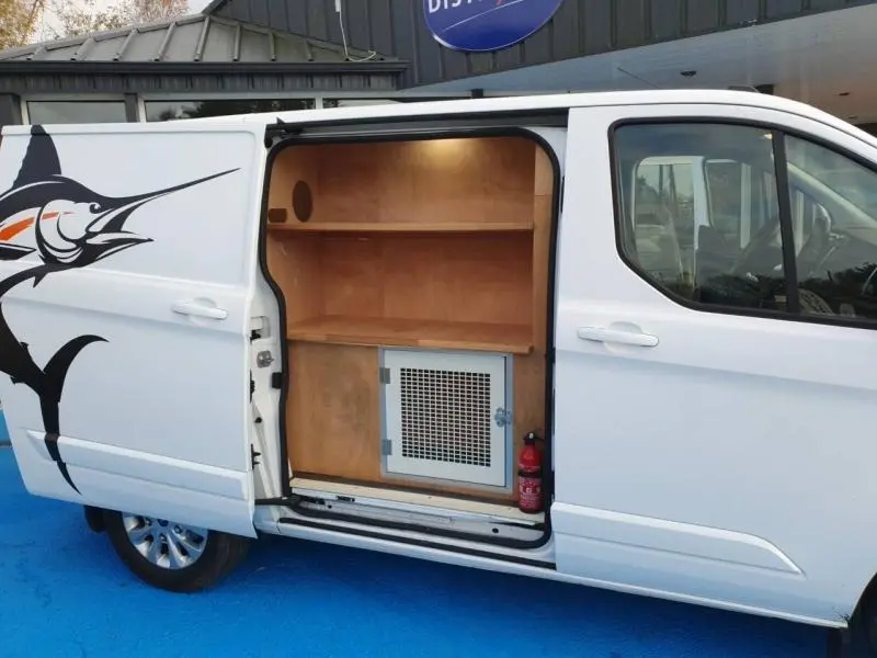 Vue latérale gauche d'un Ford Transit Custom blanc glacier avec porte coulissante ouverte révélant un aménagement en bois et un extincteur.