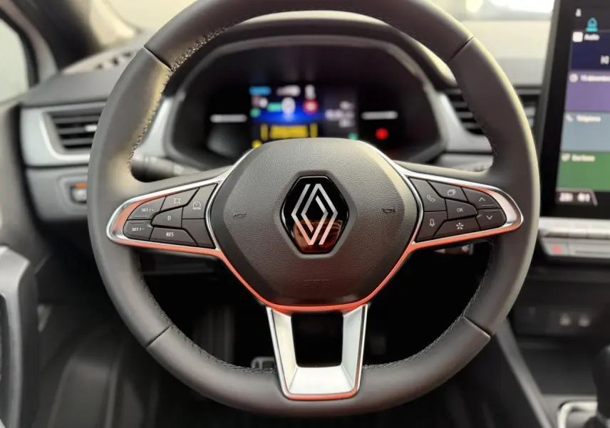 Vue rapprochée du volant en cuir noir avec commandes intégrées du Renault Captur hybride 2025, tableau de bord flou en arrière-plan.