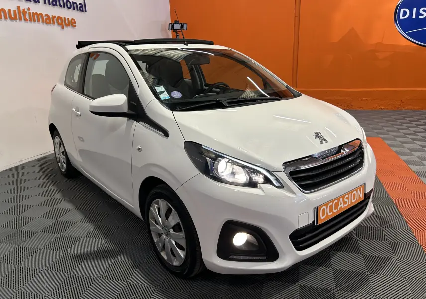 Vue 3/4 avant droite d'une Peugeot 108 blanche avec phares allumés et toit ouvrant, en showroom avec sol gris et orange.
