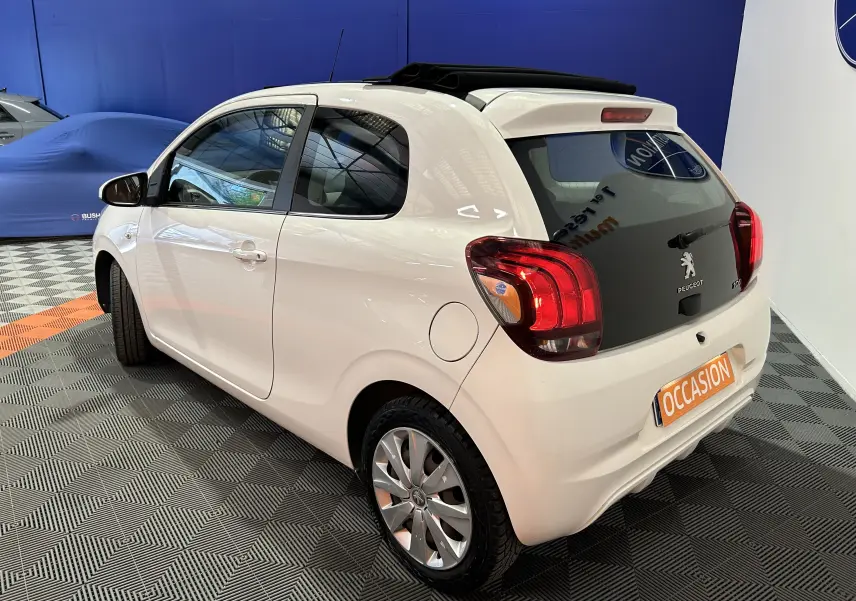 Vue 3/4 arrière droite d'une Peugeot 108 blanche avec toit ouvrant en toile, stationnée en intérieur sur sol damier.