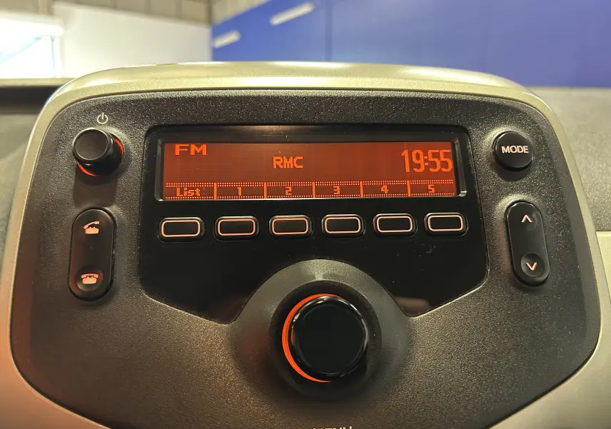 Affichage de la radio FM RMC à 19h55 sur le tableau de bord noir du Peugeot 108, détail de la console centrale.