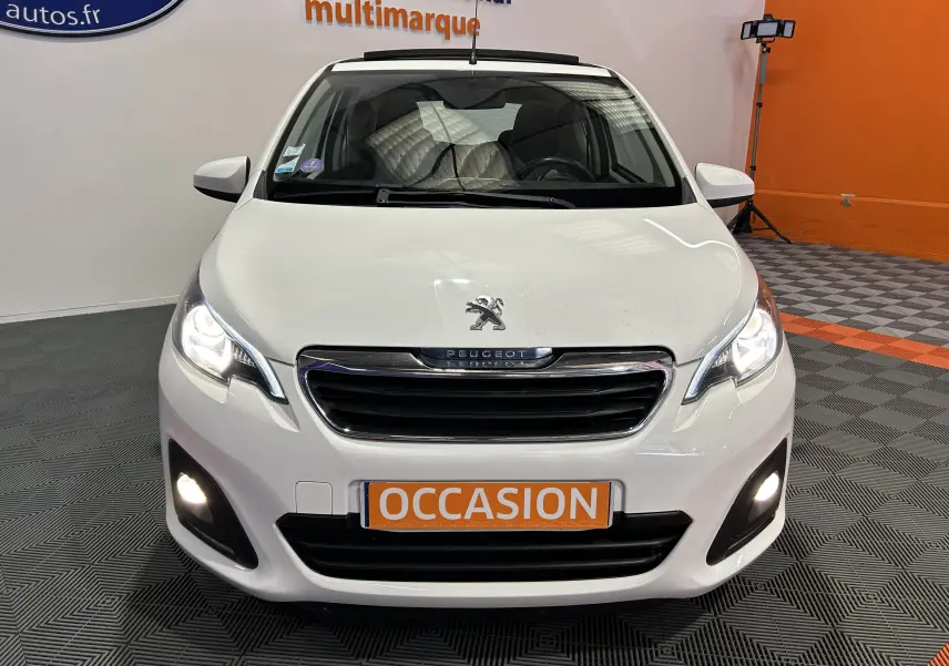 Vue avant d'une Peugeot 108 blanche avec phares et feux antibrouillard allumés dans un showroom.