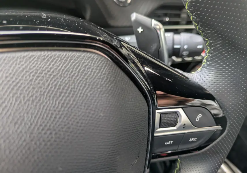 Gros plan sur le volant noir perforé avec surpiqûres vertes du Peugeot 208 Hybrid 145 e-DCS6 GT, boutons de commande visibles.