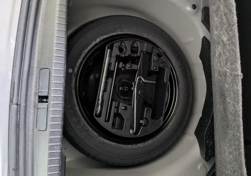 Vue rapprochée du coffre d'une Peugeot 208 Hybrid 145 gris Artense, montrant la roue de secours et les outils de bord.