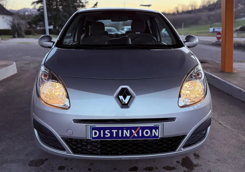 Vue avant d'une Renault Twingo II grise de 2009 avec phares allumés et plaque Distinxion visible.