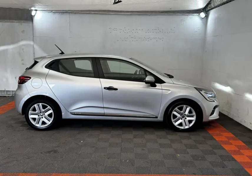 Profil droit d'une Renault Clio Business TCe 100 gris clair stationnée dans un garage avec sol noir et bandes orange.