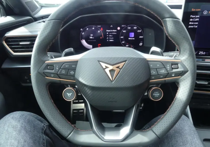 Vue rapprochée du volant perforé gris avec surpiqûres cuivrées et logo CUPRA sur le tableau de bord digital.