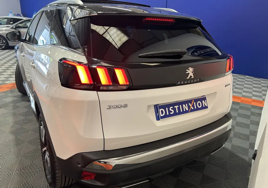 Vue 3/4 arrière droite d’un Peugeot 3008 blanc avec feux arrière allumés et toit ouvrant visible en intérieur showroom.