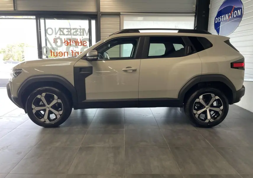 Vue de profil côté gauche d'un SUV Dacia Duster beige avec jantes alliage 18 pouces et barres de toit noires.