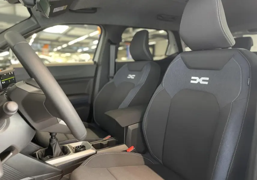 Vue intérieure avant du Dacia Duster 2025, sièges tissu Denim bleu avec logo Dacia, volant soft feel et levier de vitesses manuel.