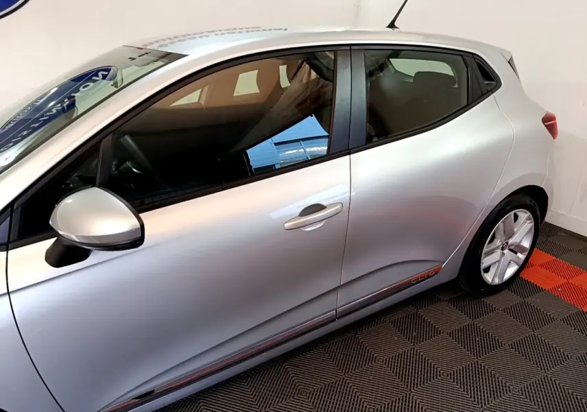 Profil droit de la Renault Clio gris clair avec poignées intégrées et jantes alliage visibles en intérieur showroom.