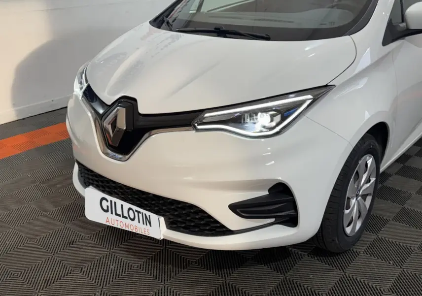Vue 3/4 avant droit d'une Renault ZOE Business blanche avec phares LED allumés et calandre noire distinctive.
