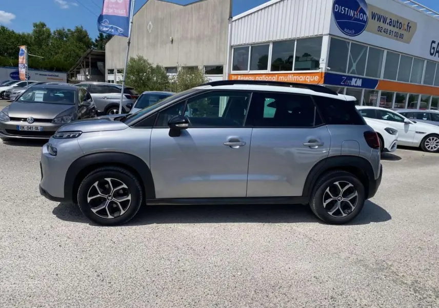Profil côté gauche d'un Citroën C3 Aircross gris Artense avec toit blanc, garé devant un concessionnaire en extérieur.