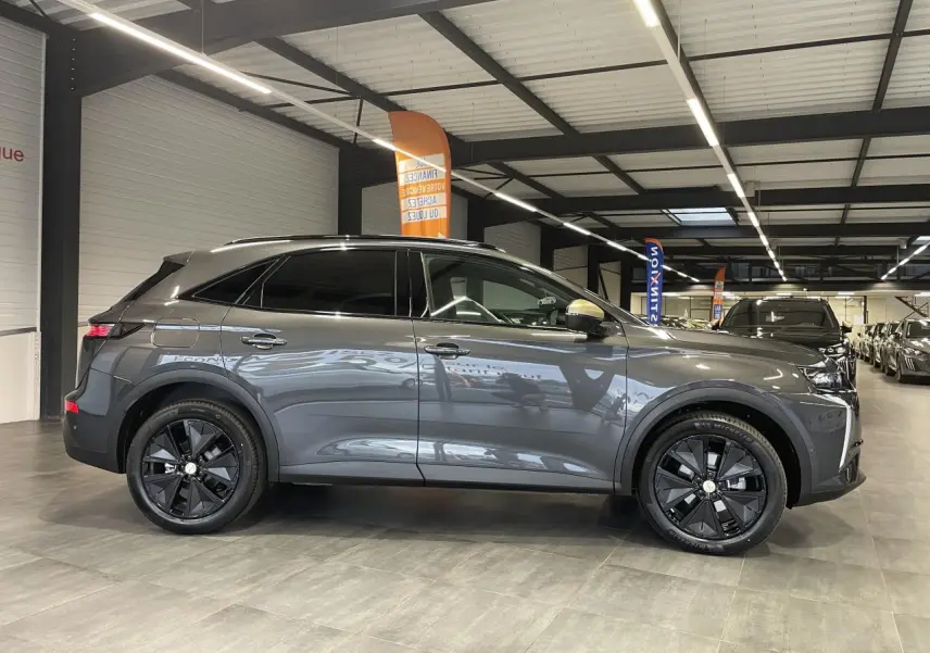 Vue de profil côté gauche du DS7 Crossback gris titanium avec jantes noires et barres de toit noir brillant en intérieur showroom.