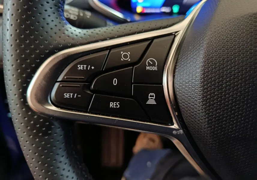 Gros plan sur les commandes noires du volant perforé du Renault Symbioz 2025, version hybride bleu visible en arrière-plan.