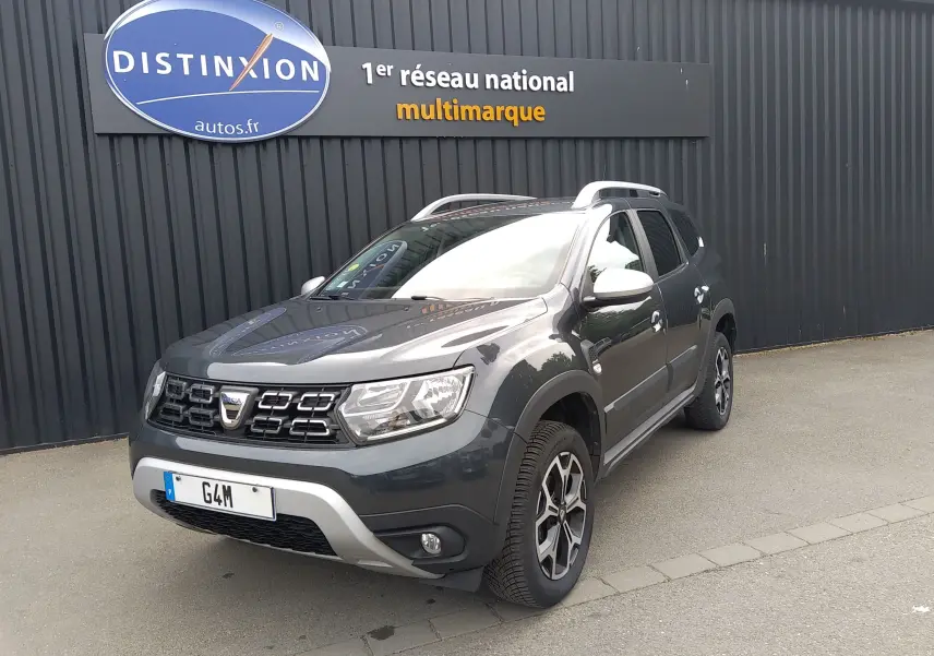 Dacia Duster gris 3/4 avant droit avec jantes alliage et barres de toit, devant un mur noir avec enseigne Distinxion.