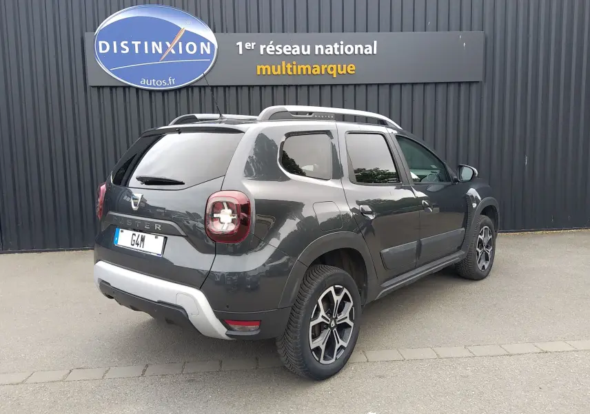 Vue 3/4 arrière droite d'un Dacia Duster gris 2020 avec jantes alliage et barres de toit, stationné devant un mur noir.