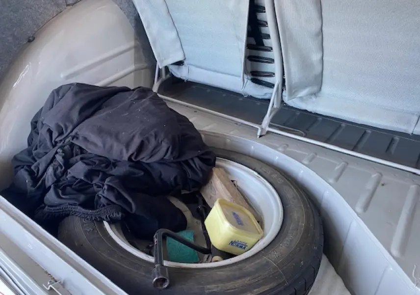 Coffre ouvert d'une Citroën 2 CV beige clair de 1968, montrant une roue de secours et des objets divers à l'intérieur.