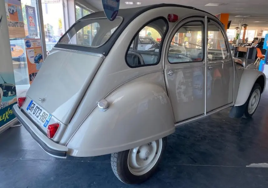Vue 3/4 arrière droite d'une Citroën 2 CV beige clair refaite à neuf, exposée en intérieur avec ses phares arrière rouges.