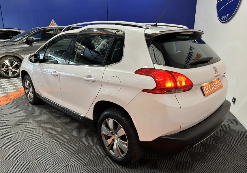 Peugeot 2008 blanc vue 3/4 arrière droit, feux arrière allumés et jantes alliage visibles en intérieur showroom.