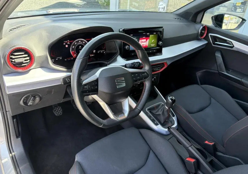 Vue intérieure côté conducteur du SEAT Arona 2024, volant sport cuir noir avec surpiqûres rouges et tableau de bord digital.