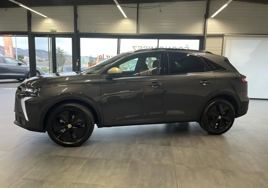 Vue de profil côté gauche du DS7 Crossback gris titanium avec jantes noires et coques de rétroviseurs dorées satinées en intérieur showroom.