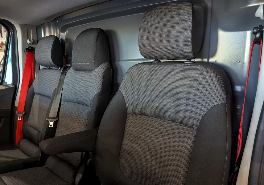 Vue intérieure rapprochée des sièges avant en tissu gris foncé du Renault Trafic Fourgon L2H1 2025 avec ceintures rouges.