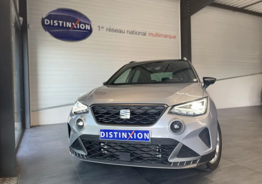 Vue avant d'une SEAT Arona 2024 gris clair avec calandre noire et phares allumés en intérieur showroom.