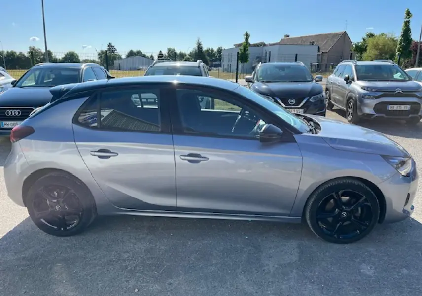 Profil côté gauche d'une Opel Corsa gris métallisé avec toit noir et jantes noires, stationnée en extérieur au soleil.