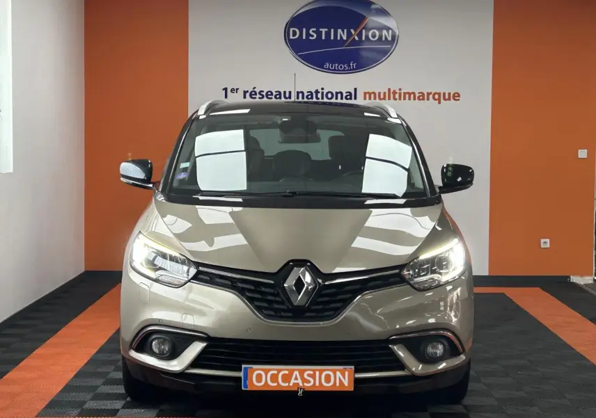 Vue frontale d'un Renault Scénic Grand IV marron clair avec feux allumés dans un showroom orange et noir.