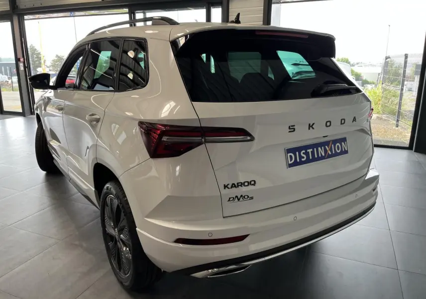 Vue 3/4 arrière droite d'un SUV SKODA KAROQ blanc 2025 avec jantes noires et hayon électrique en intérieur showroom.