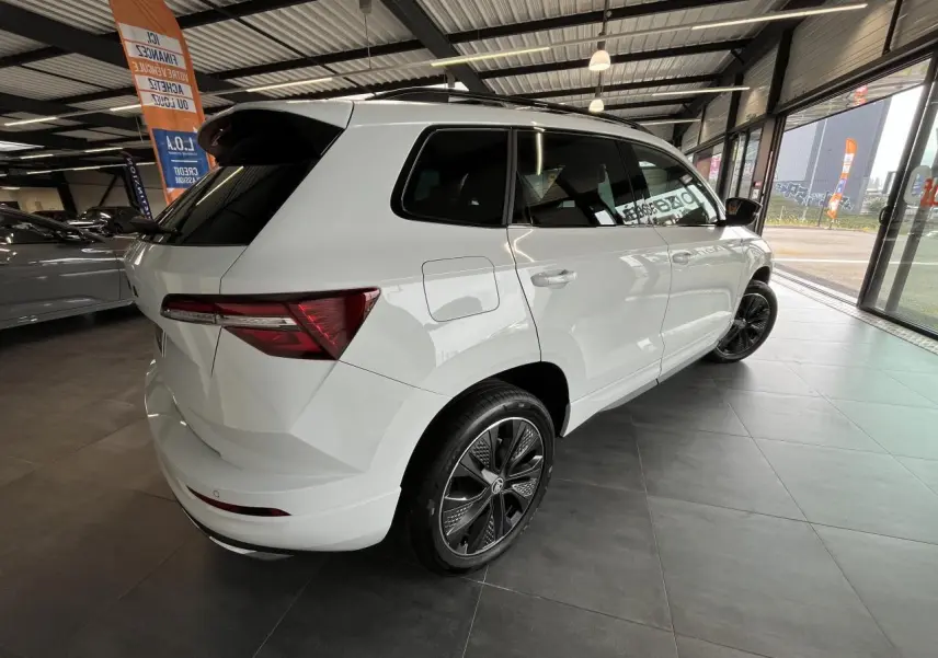 Vue 3/4 arrière droite d'un SUV Skoda Karoq blanc 2025 avec jantes alliage noires et vitres surteintées en showroom.