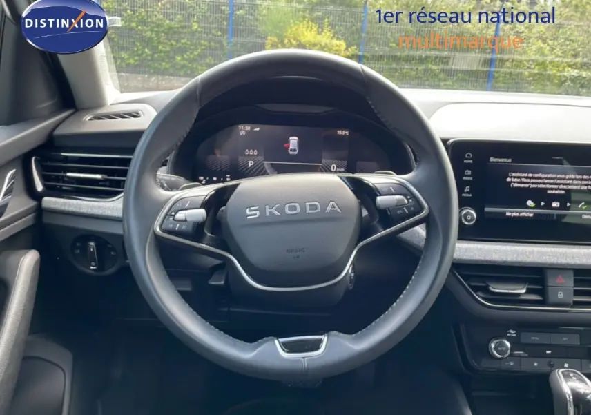 Vue intérieure centrée sur le volant noir SKODA avec tableau de bord digital et écran tactile sur fond gris.