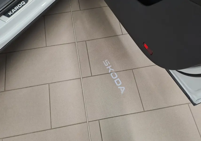 Projection lumineuse du logo SKODA au sol, vue partielle de la porte ouverte d'un Karoq blanc Lune.
