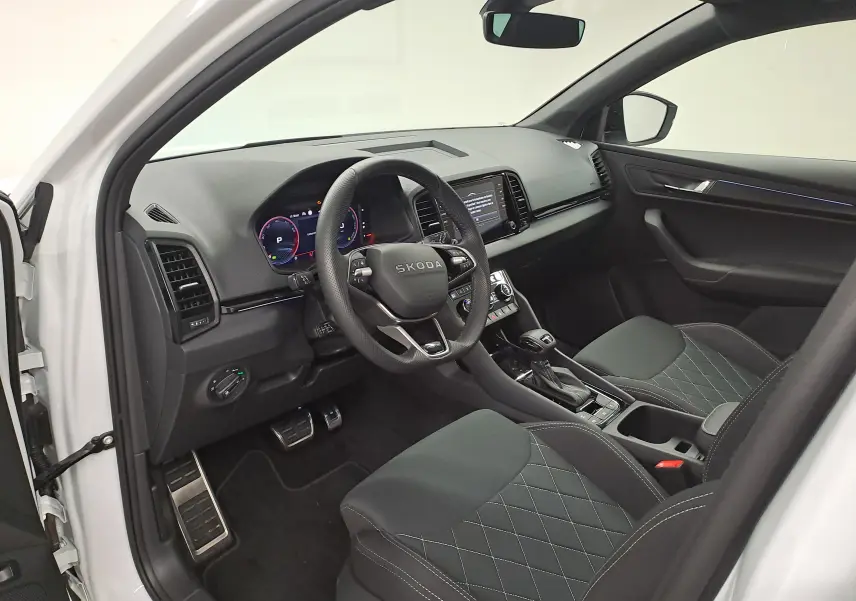 Vue intérieure côté conducteur du Skoda Karoq blanc lune 2025, avec volant multifonction et sièges en tissu noir matelassé.