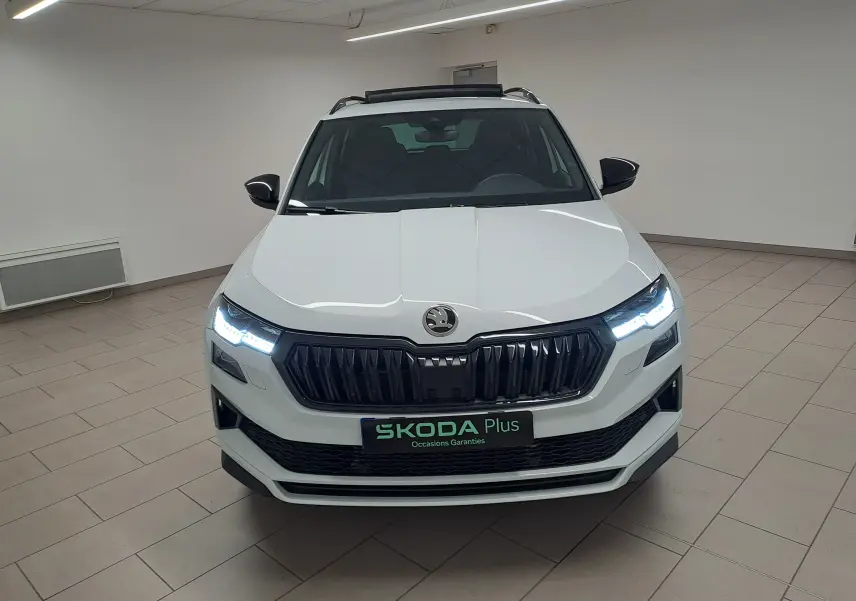 Vue avant d'un Skoda Karoq blanc lune 2025 avec phares allumés et toit ouvrant panoramique visible en intérieur.