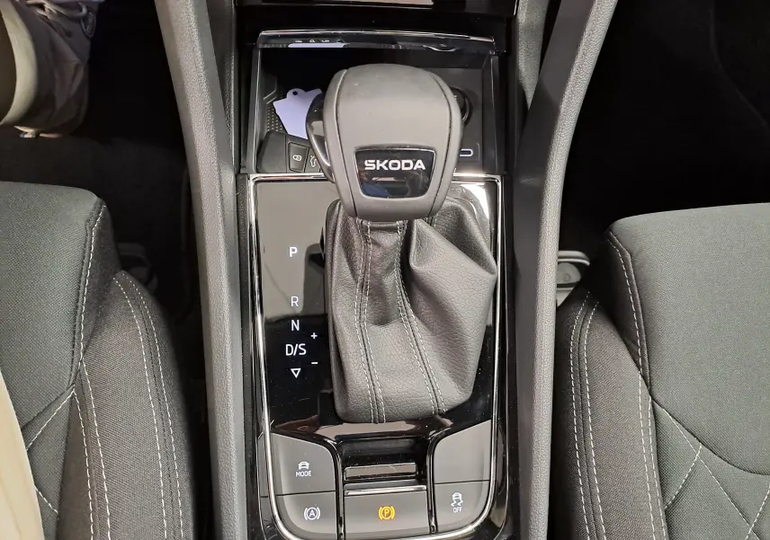 Vue plongeante sur le levier de vitesse automatique noir marqué SKODA, entouré de sièges tissu gris avec surpiqûres blanches.