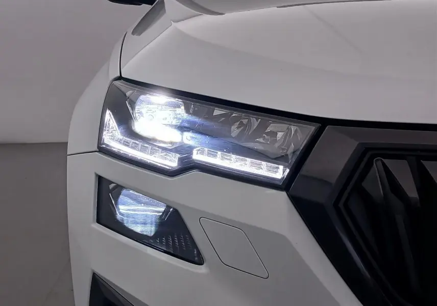 Gros plan sur le phare avant gauche allumé d’un Skoda Karoq blanc lune 2025 avec calandre noire sportive.