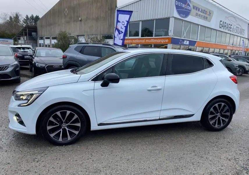 Profil côté gauche d'une Renault Clio blanche 2023 avec jantes alliage noires et vitres surteintées, stationnée en extérieur.
