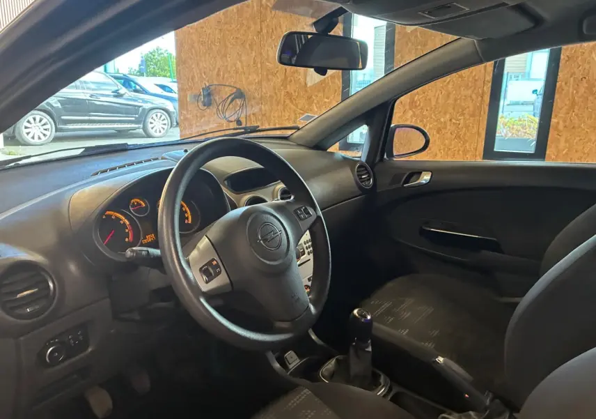 Vue intérieure côté conducteur de l’Opel Corsa gris clair 2012, volant multifonction et sellerie tissu noir visibles.