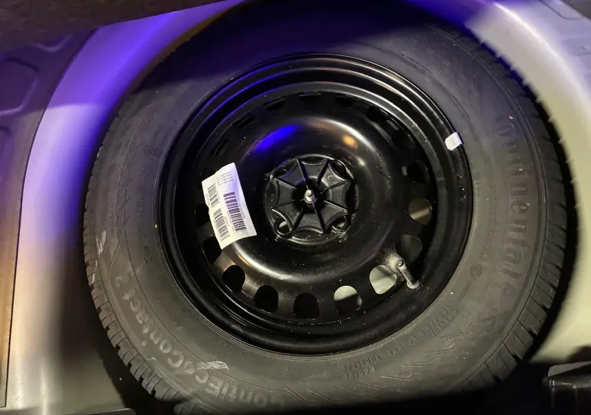 Gros plan sur la roue de secours noire d'une Opel Corsa gris clair, visible dans le coffre sous un éclairage tamisé.