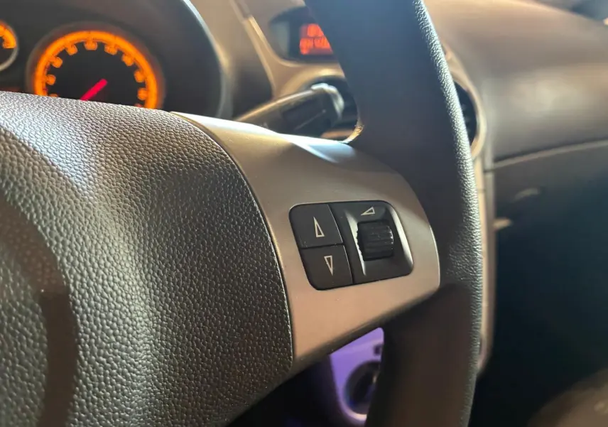 Gros plan sur le volant noir de l’Opel Corsa gris clair 2012, avec commandes intégrées et tableau de bord éclairé en orange.