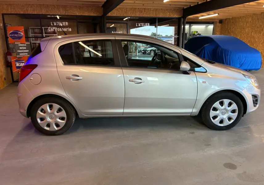 Vue de profil côté gauche d'une Opel Corsa gris clair 5 portes de 2012 stationnée en intérieur, avec jantes acier et enjoliveurs.